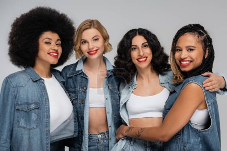 Gruppe multikultureller Smiley-Freundinnen in Jeanskleidung blickt in die Kamera auf grau, Vielfalt