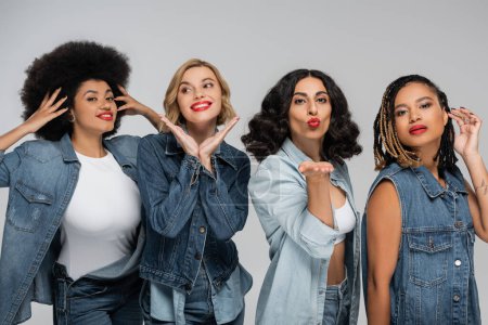 multiracial woman blowing air kiss near happy charming girlfriends posing in denim clothes on grey
