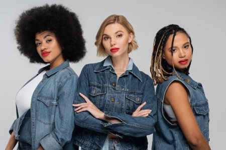 blonde woman with folded arms near trendy african american girlfriends in denim clothes on grey