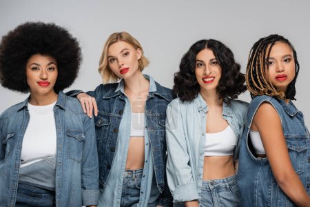 diverse multiracial women in blue denim outfit looking at camera on grey, group fashion portrait