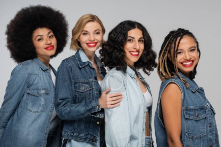 joyful multicultural female models in casual denim clothes smiling at camera on grey, diverse beauty