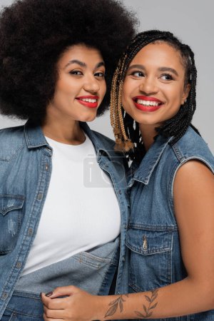 tätowierte und fröhliche afrikanisch-amerikanische Frau in Jeanskleidung lächelt neben trendiger Freundin auf grau