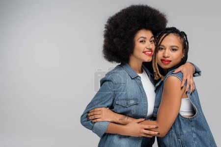 fröhliche und trendige afrikanisch-amerikanische Freundinnen, die sich vor grauem Hintergrund umarmen, Jeansmode