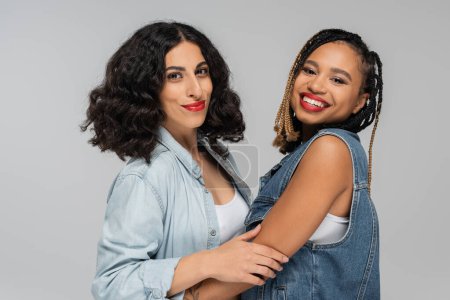 zwei unbeschwerte multirassische Freundinnen in stylischer Jeanskleidung, die in grau und zu zweit in die Kamera lächeln