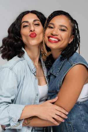 Photo for Multiracial woman pouting lips near cheerful african american girlfriend in blue denim wear on grey - Royalty Free Image