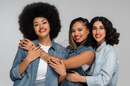 stylische und Smiley multiethnische Freundinnen in blauen Jeans tragen Blick in die Kamera auf grau, Vielfalt