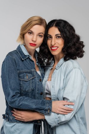 Photo for Diverse beauty, happy multiracial female friends in blue denim attire embracing on grey backdrop - Royalty Free Image