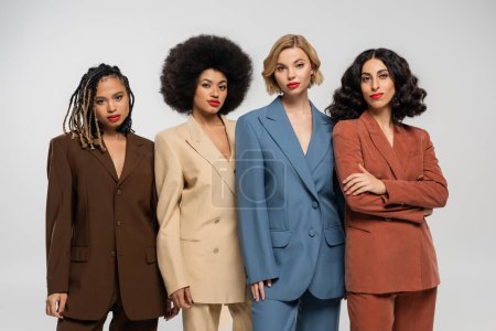 group of elegant multiethnic women in colorful suits looking at camera on grey, diverse girlfriends
