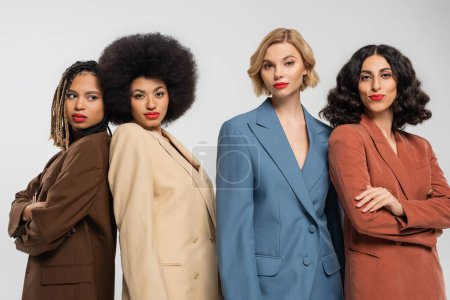 group of elegant multiethnic women in colorful suits looking at camera on grey, trendy girlfriends