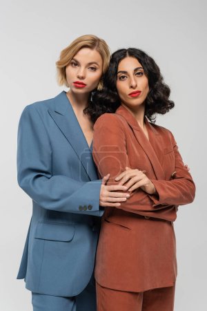 stylish blonde woman embracing brunette multiracial girlfriend on grey, models in colorful suits