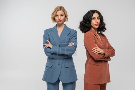 multiethnic blonde and brunette female models in colorful suits posing with folded arms on grey