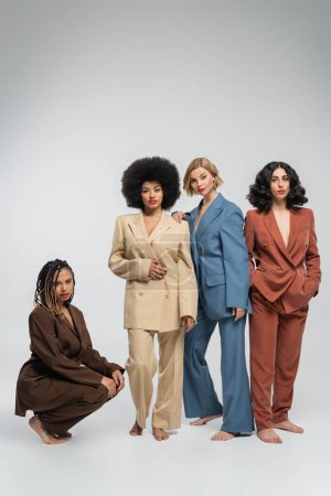 full length of multiracial barefoot girlfriends in colorful suits on grey backdrop, diverse beauty