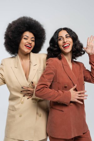Téléchargez les photos : Joyeuse femme multiraciale agitant la main près de petite amie afro-américaine sur gris, costumes colorés - en image libre de droit