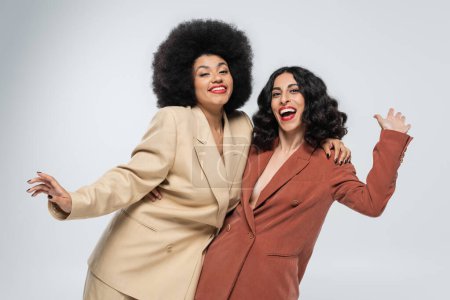 Photo for Carefree multiethnic girlfriends in trendy colorful suits waving hands on grey, fashion and joy - Royalty Free Image
