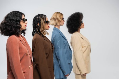 Photo for Side view of stylish multiracial girlfriends in dark sunglasses and multicolored suits on grey - Royalty Free Image