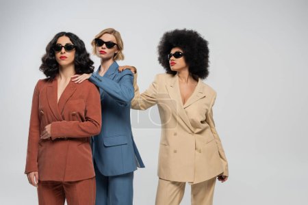 multiethnic girlfriends in colorful suits and sunglasses touching shoulders of each other on grey