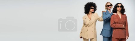 Téléchargez les photos : Copines multiraciales en costumes et lunettes de soleil se touchant les épaules sur gris, bannière - en image libre de droit
