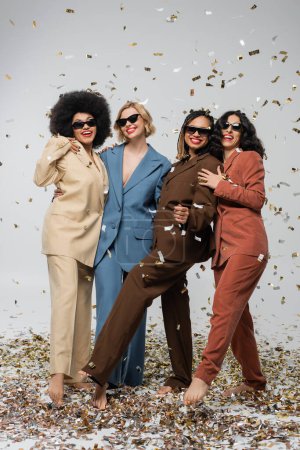 Unbekümmerte multiethnische Freundinnen in Sonnenbrillen und Anzügen neben buntem Konfetti auf grau, Party-Time