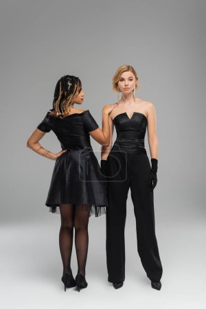 Photo for Elegant african american woman touching shoulder of stylish blonde girlfriend on grey, fashion shoot - Royalty Free Image