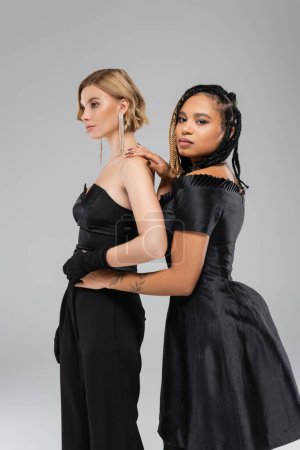 Photo for Elegant african american woman in black attire embracing waist of blonde glamour friend on grey - Royalty Free Image