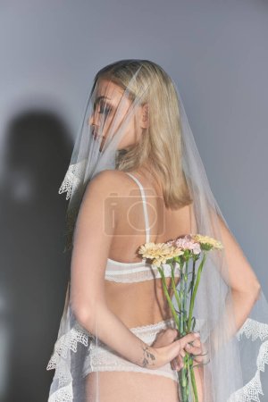 attraktive sexy Frau in weißen Dessous mit Schleier hält Blumen hinter ihrem Rücken auf grauem Hintergrund