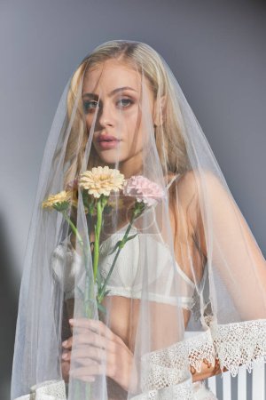 vertical shot of attractive young woman wearing veil posing with flowers in hands looking ta camera