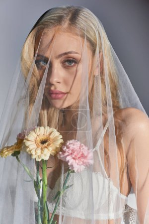 vertical shot of sexy woman with blonde hair in white veil holding flowers and looking at camera