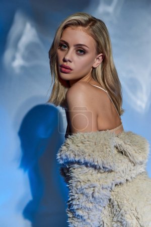 vertical shot of attractive woman with blonde hair in faux fur looking at camera on blue backdrop