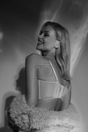 Schwarz-Weiß-Aufnahme einer fröhlich verführerischen Frau in sexy Dessous und Kunstpelz, die den Kopf leicht dreht