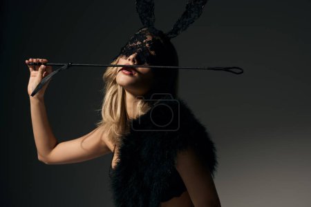 appealing young woman with blonde hair in rabbit mask posing with bdsm whip and looking at camera