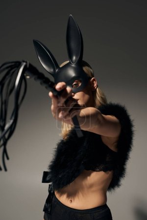 vertical shot of alluring woman in sexy lingerie with rabbit mask pointing bdsm whip at camera