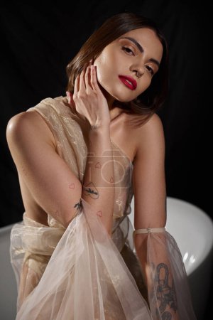 pretty woman with tattoos posing in transparent dress posing near bathtub with black backdrop