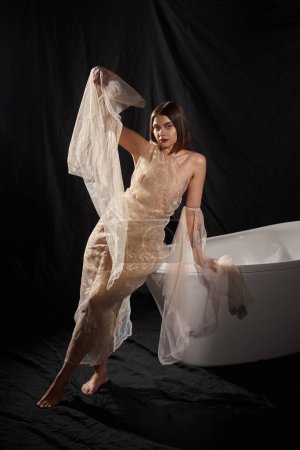 full length, barefoot woman posing in transparent dress posing near bathtub with black backdrop