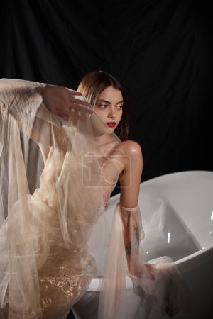 young woman with short hair posing in transparent dress posing near bathtub with black backdrop