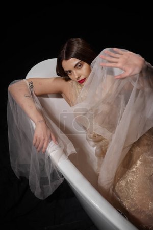 high angle view of graceful model in transparent dress gesturing while lying on bathtub on black