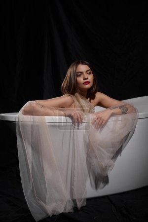 young graceful model in transparent dress while sitting inside of bathtub on black backdrop