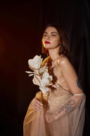 young and graceful woman in transparent dress holding white blooming flower on black backdrop