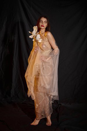 full length of graceful woman in transparent dress holding white blooming flower on black backdrop
