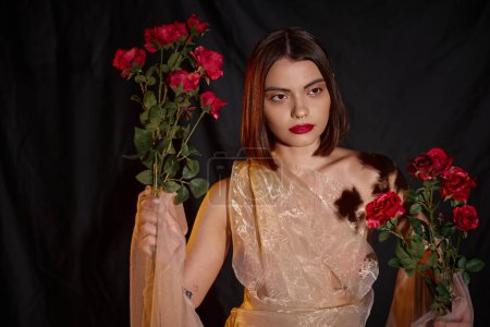 charming young woman in romantic transparent dress holding blooming red roses on black backdrop