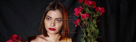 charming young woman with red lips holding blooming red roses on black backdrop, banner