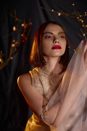 sensual young woman in transparent dress posing near blooming flowers on branches on black backdrop