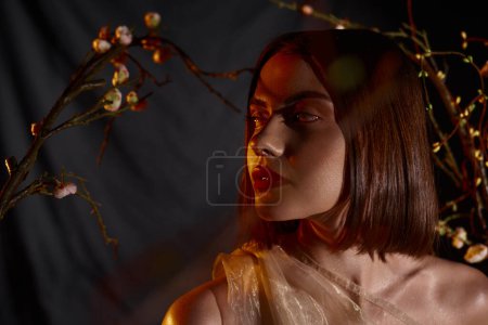 dreamy young woman in transparent dress posing near blooming flowers on branches on black backdrop