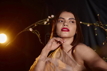 delicate young woman in transparent dress posing near blooming flowers on branches on black backdrop