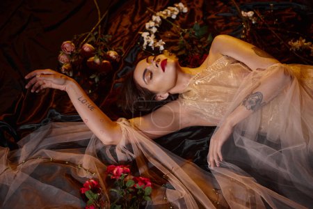 mujer joven encantadora y tatuada en vestido transparente romántico acostado entre flores en flor