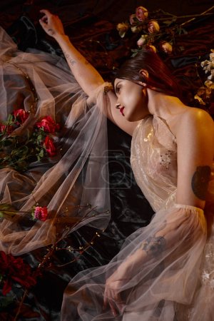 schöne und tätowierte junge Frau in romantischem transparentem Kleid zwischen blühenden Blumen liegend