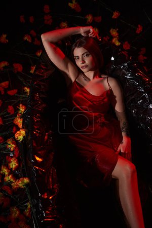 top view, sensual young woman in red slip dress lying in black bathtub among beautiful flowers
