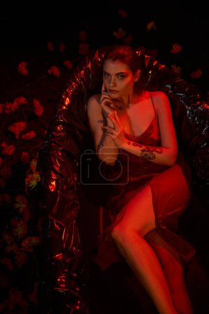 Photo for Top view, young sensual woman in red slip dress lying in black bathtub among beautiful flowers - Royalty Free Image