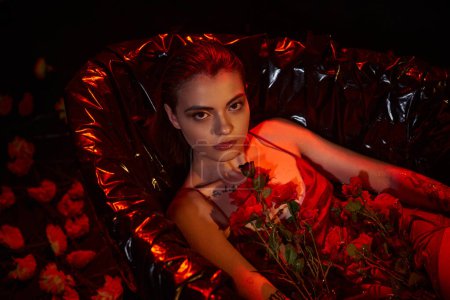 attractive young woman in wet slip dress lying in black bathtub with blooming flowers, red light