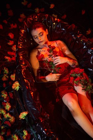 pretty young woman in wet slip dress lying in black bathtub with blooming flowers, red light