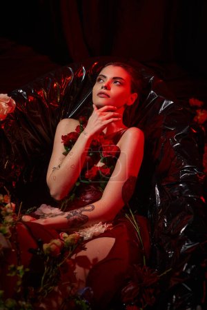 attractive young woman in wet slip dress sitting in black bathtub with blooming flowers, looking up